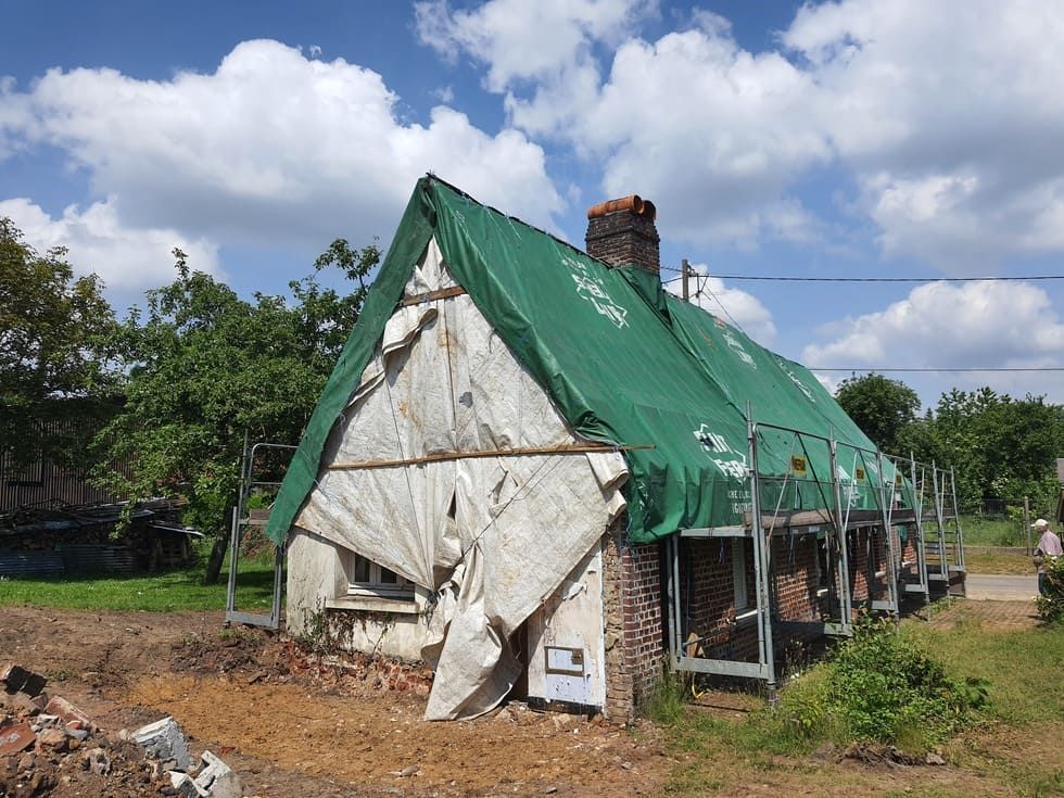 Maisonnette en rénovation, recouverte d'une bâche verte, échafaudages, cheminée en briques, débris au premier plan, journée ensoleillée.