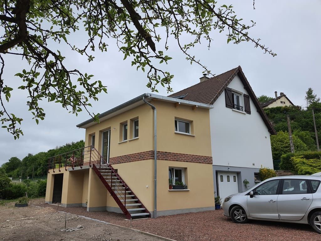 Maison à deux étages avec façade jaune et blanche, escalier et voiture argentée garée dans l'allée.