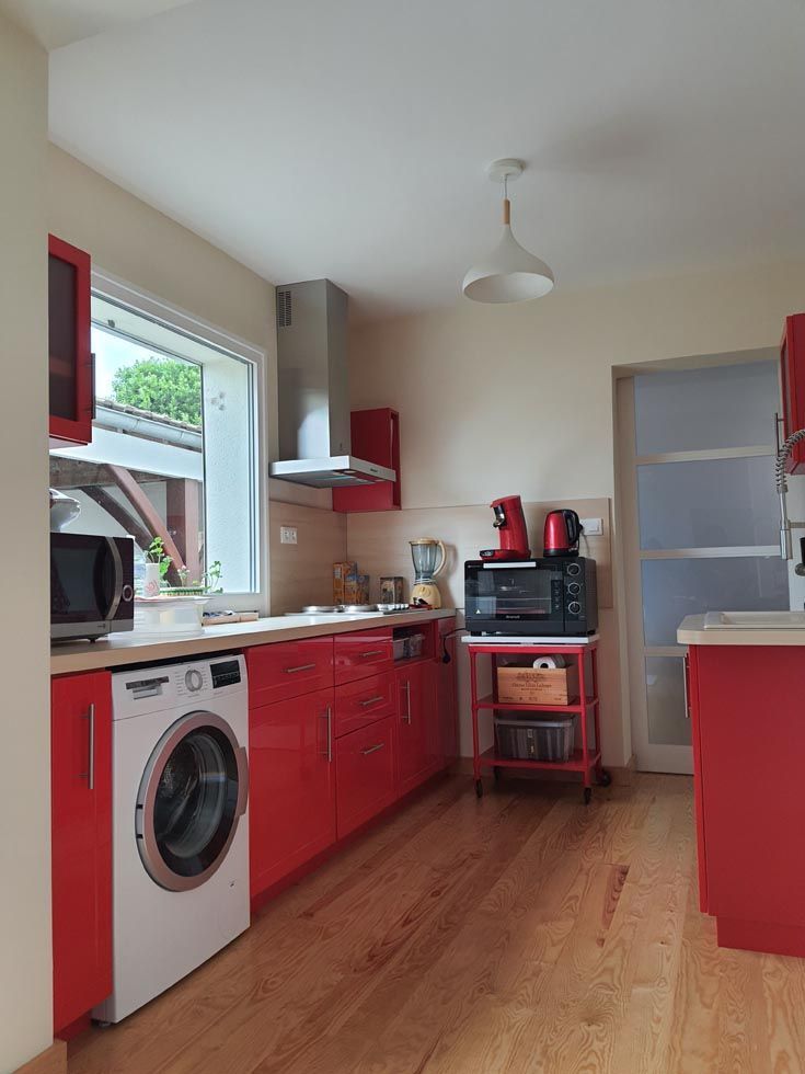Cuisine rouge avec placards, électroménager et lave-linge. Fenêtre, luminaire et parquet.