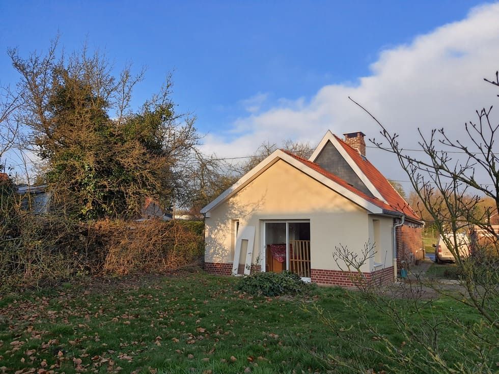 Petite maison de plain-pied avec des éléments en briques rouges et une cheminée. Par une journée ensoleillée, la végétation luxuriante entoure le bâtiment.