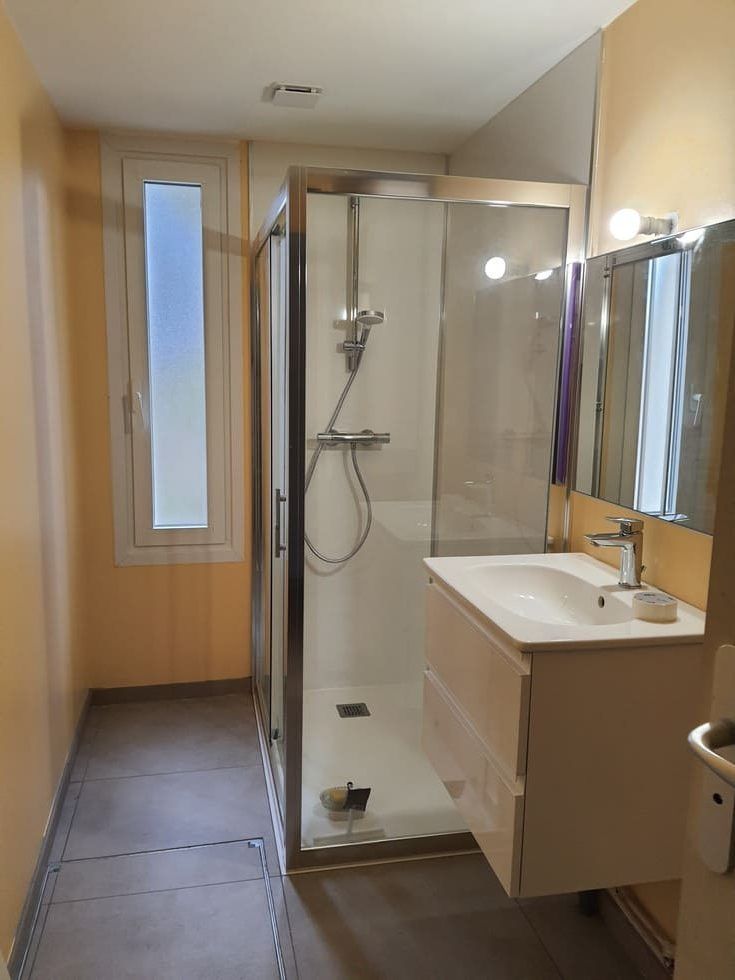 Salle de bains avec cabine de douche, meuble-lavabo, petite fenêtre, sol gris, murs beiges.