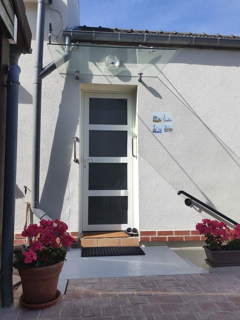 Porte blanche avec auvent en verre, paillasson noir, fleurs roses en pot et allée de briques.