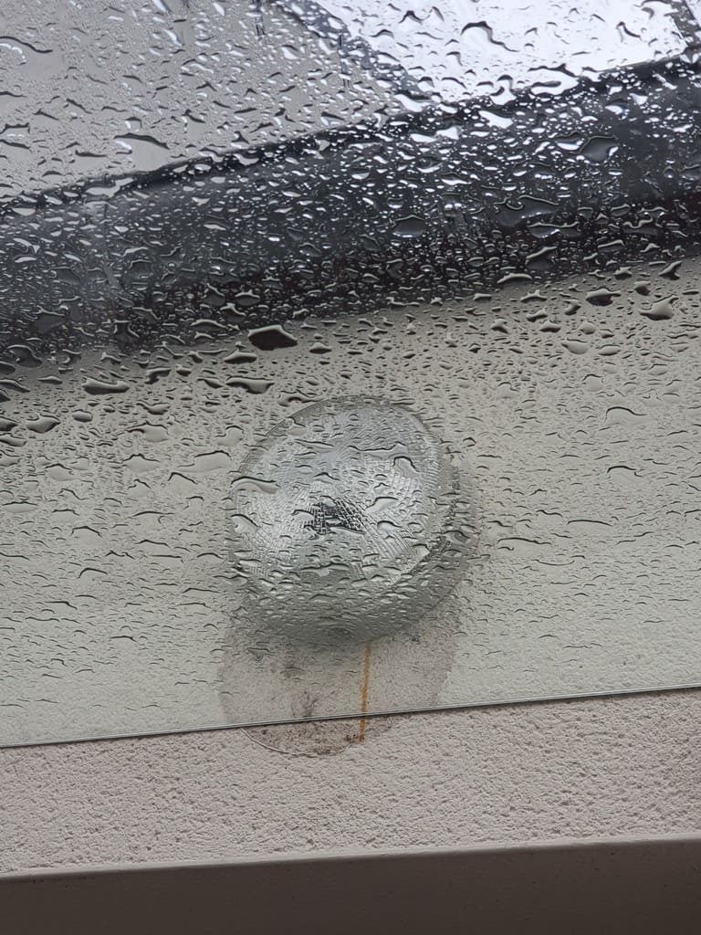 Grille d'aération recouverte de gouttelettes d'eau sur un mur blanc, sous un toit ruisselant de pluie.