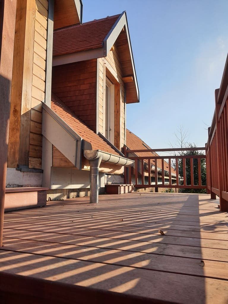 Terrasse en bois ombragée, à côté d'un bâtiment au toit et au bardage rouges, par une journée ensoleillée.