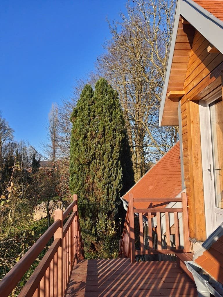 Balcon en bois avec rambarde, donnant sur un conifère verdoyant et des arbres se détachant sur un ciel bleu, à côté d'une structure ressemblant à un chalet.