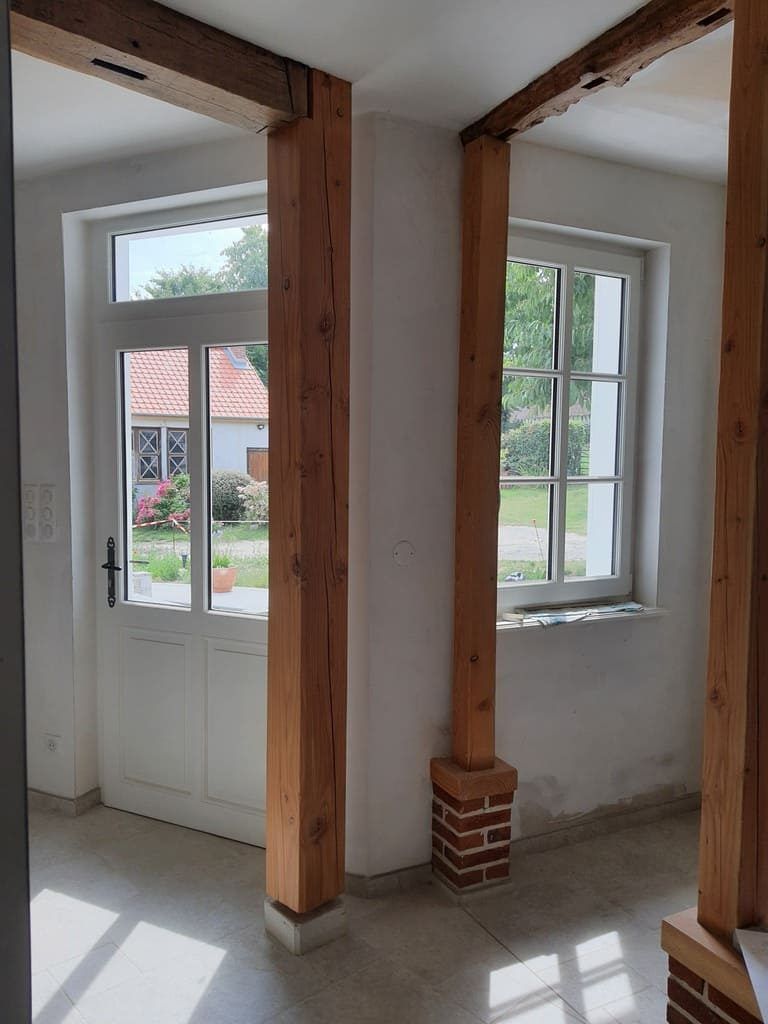 Intérieur avec poutres et colonnes en bois apparentes, fenêtres et porte donnant sur le jardin extérieur.