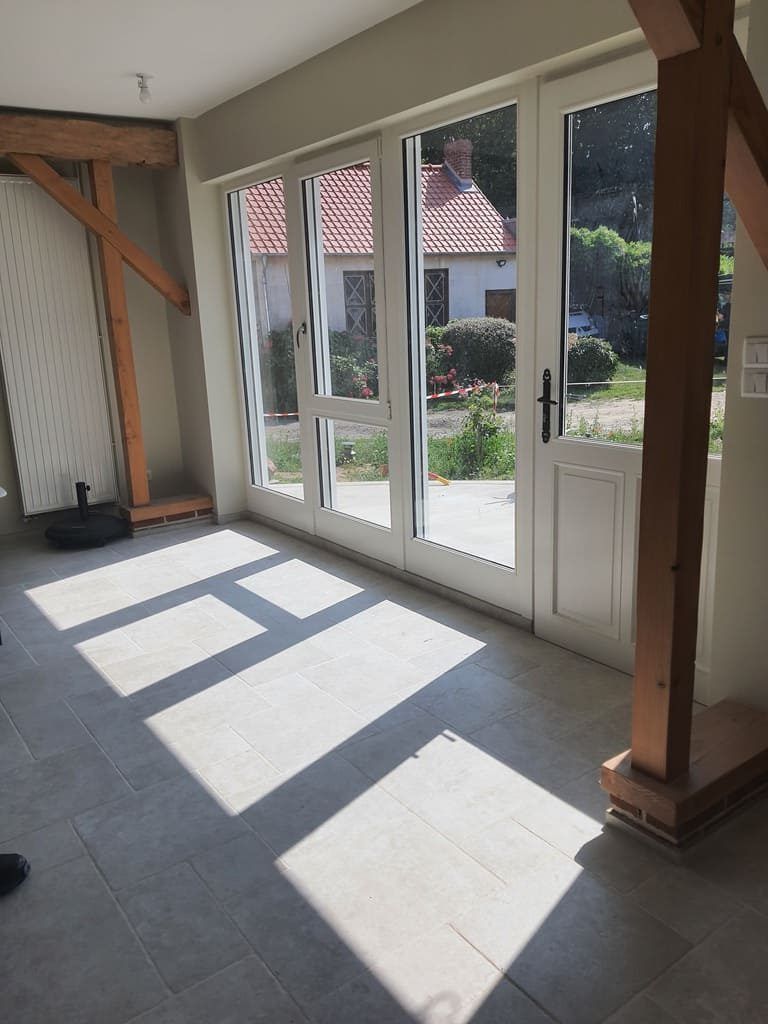 Intérieur baigné de lumière naturelle grâce à ses grandes fenêtres et ses poutres apparentes. On aperçoit une maison au loin à travers les fenêtres.