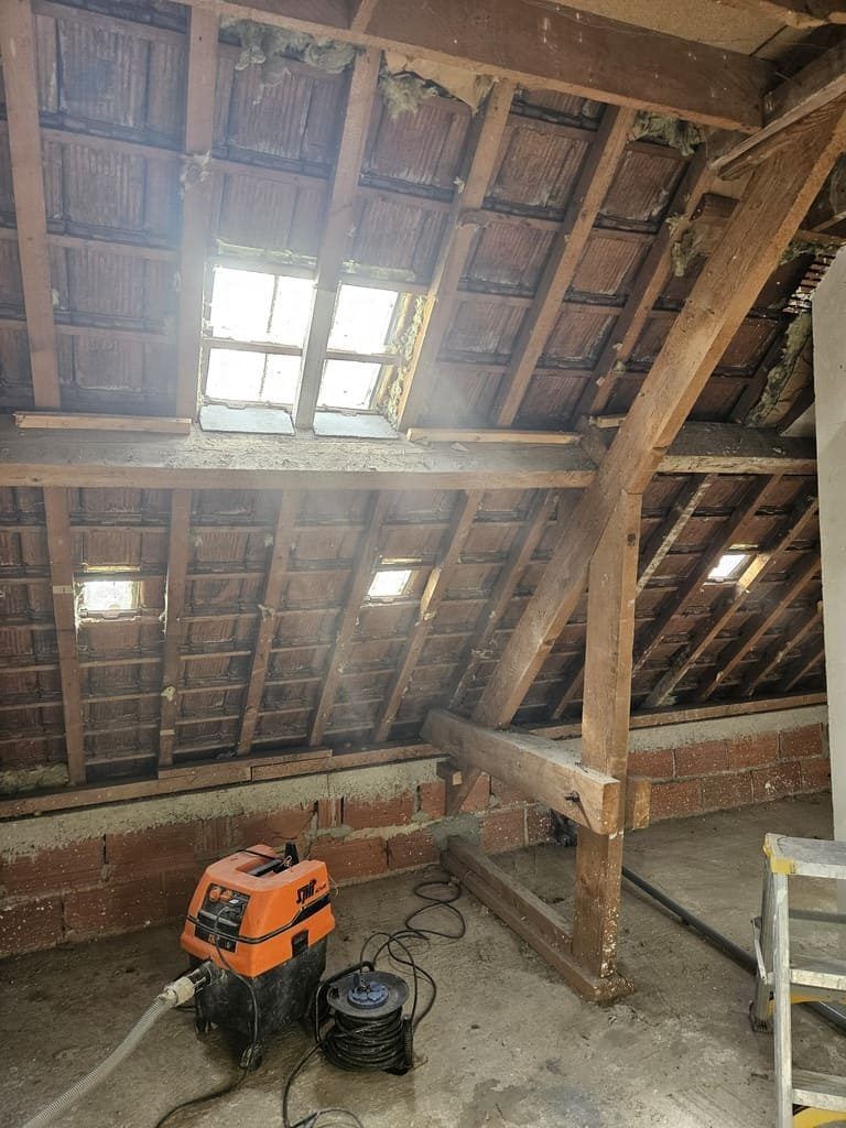 Vue intérieure d'un grenier avec poutres apparentes, tuiles et un aspirateur au sol.