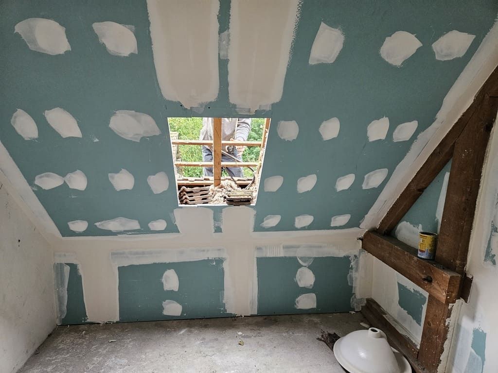 Pièce aux murs bleus, pose de cloisons sèches en cours, poutres en bois apparentes et puits de lumière.