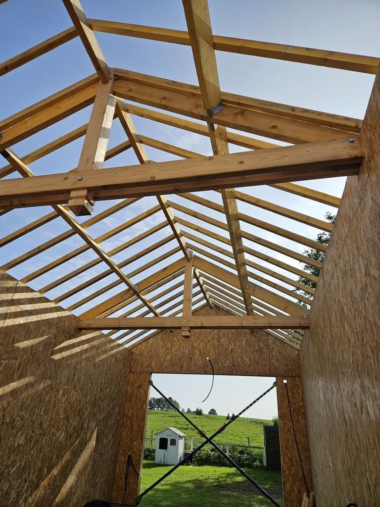 Vue intérieure d'une structure en bois en construction, avec poutres apparentes et murs en panneaux OSB, sur fond de ciel bleu.