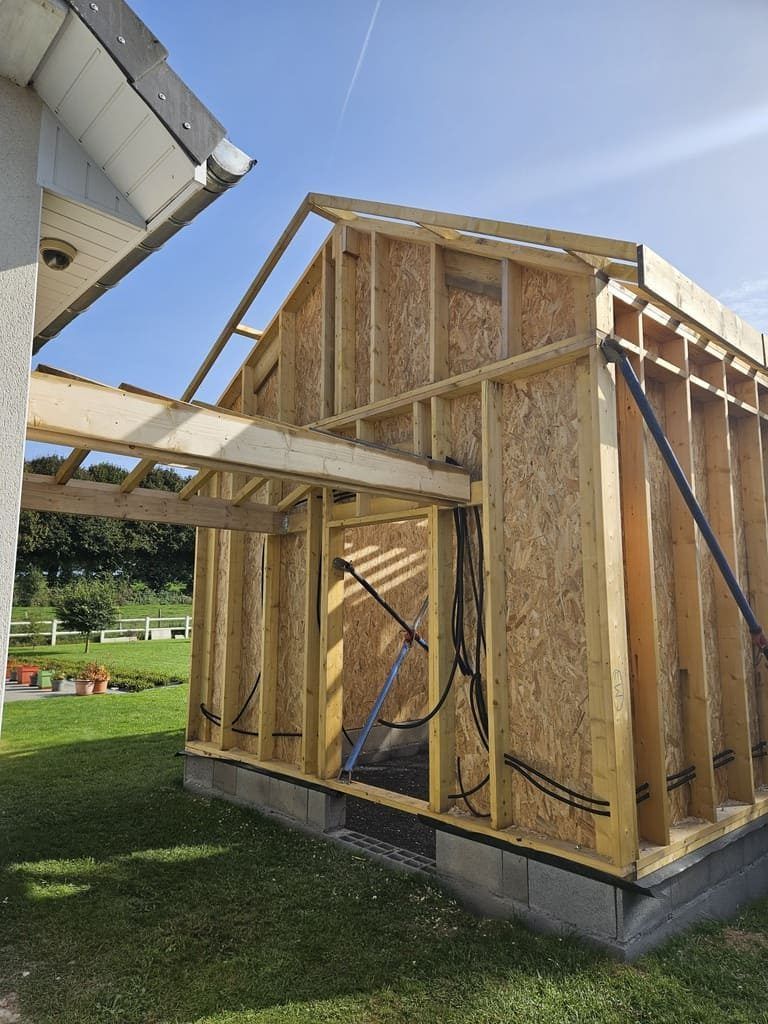 Construction d'une structure à ossature bois, adossée à une maison, sur des fondations en béton, en extérieur sous un ciel bleu.