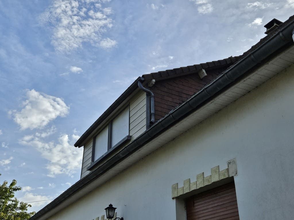 Maison avec lucarnes et toit de tuiles rouges sur un ciel partiellement nuageux.