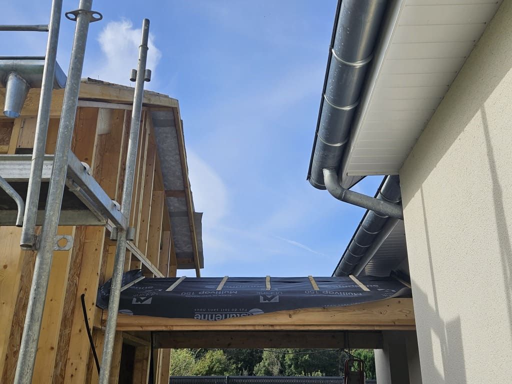 Construction de deux bâtiments : l’un à ossature bois, l’autre avec une gouttière. On aperçoit un ciel bleu au-dessus.