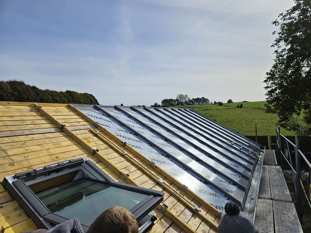Toiture avec panneaux métalliques partiellement installés, puits de lumière et échafaudages sur fond de champ verdoyant sous un ciel bleu.