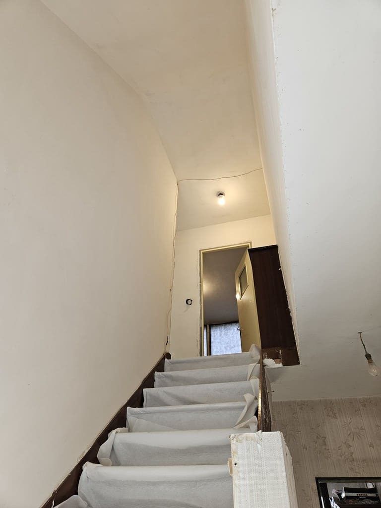 Escalier montant avec des marches recouvertes de blanc, des murs blancs et une lumière au palier supérieur.
