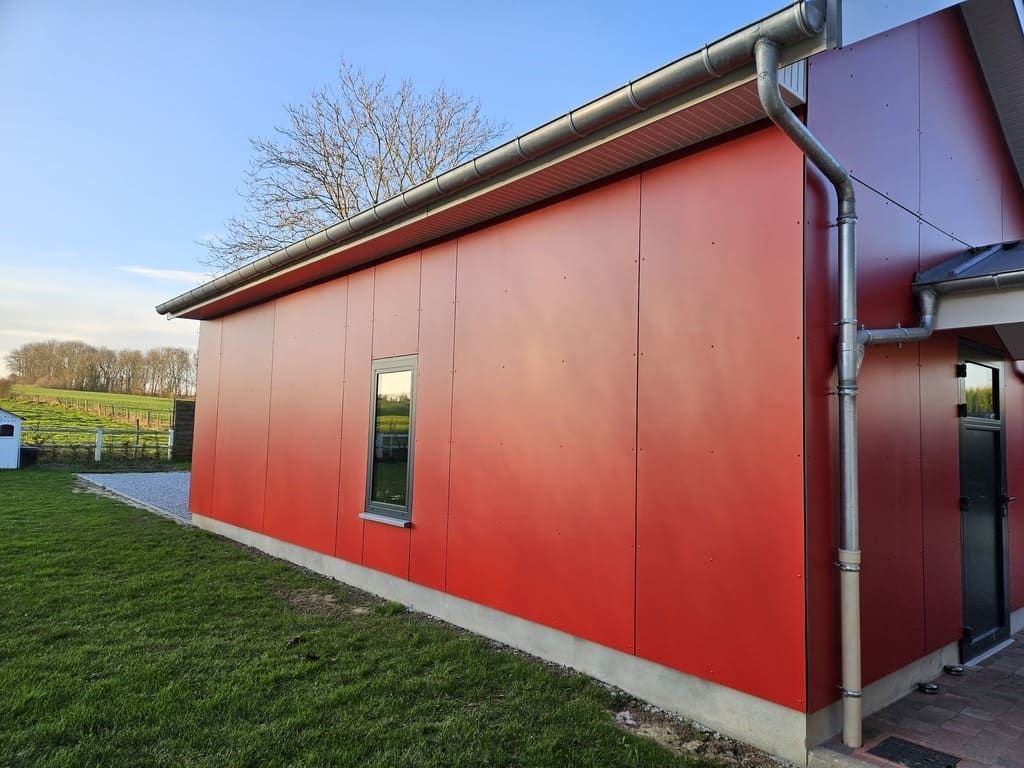 Bâtiment à façade rouge avec une petite fenêtre, une gouttière et un toit en métal, se détachant sur un jardin herbeux et un ciel nuageux.