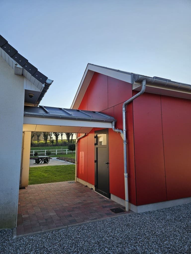 Un hangar rouge à porte noire, relié à un bâtiment blanc par une allée couverte et un pavage en briques.
