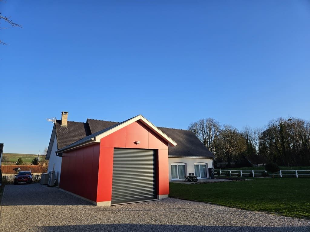 Garage rouge devant une maison, allée de gravier. Ciel bleu au-dessus.