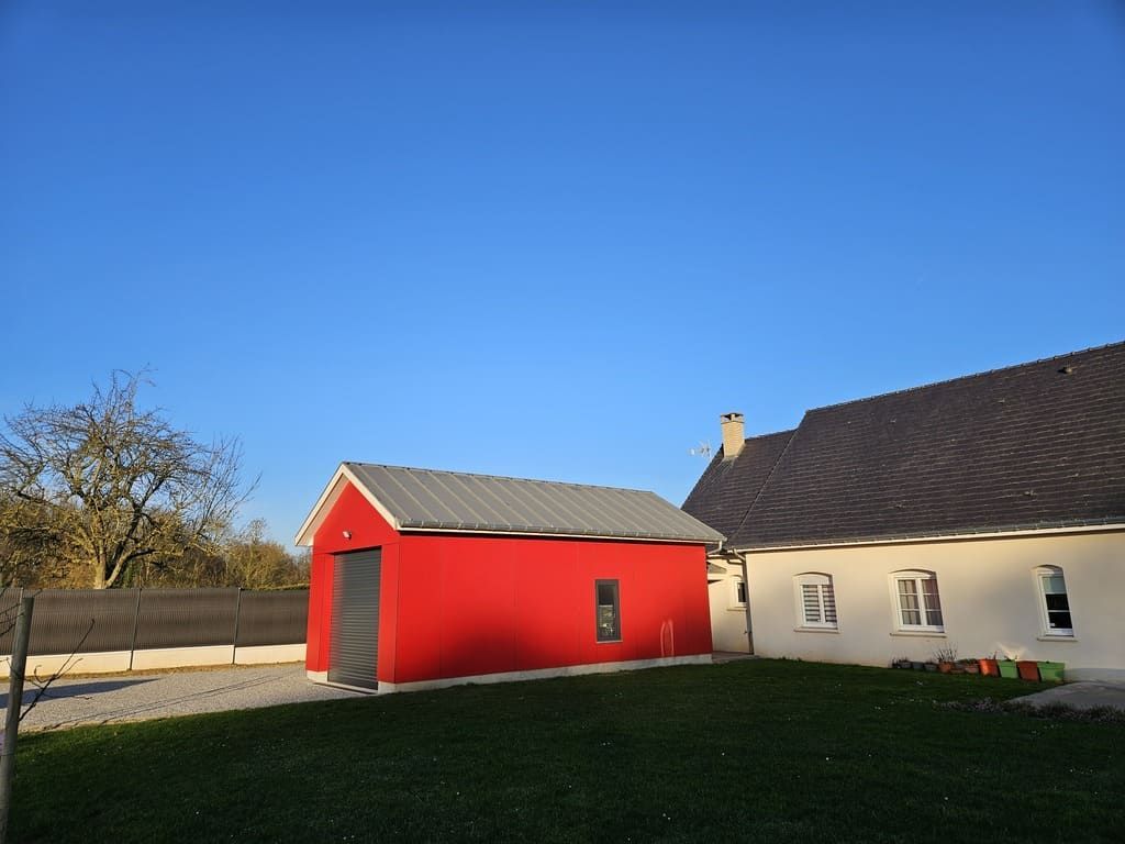Un hangar rouge vif au toit gris se dresse à côté d'une maison blanche sous un ciel d'un bleu limpide.