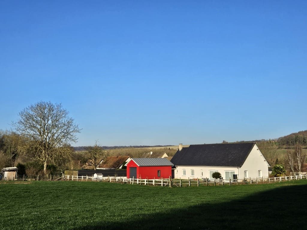 Un champ verdoyant, des maisons aux murs blancs et rouges, et un arbre sous un ciel bleu limpide.