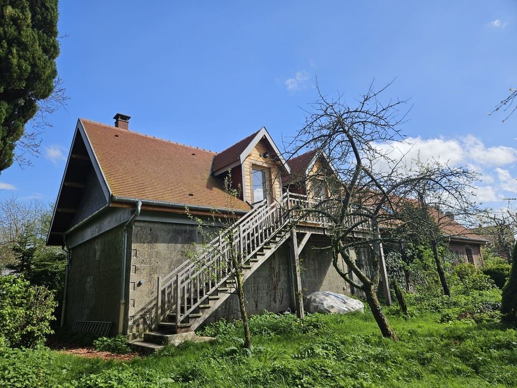 Maison à deux étages avec escalier extérieur, toit rouge, murs en béton, entourée d'arbres et d'herbe, sous un ciel bleu.
