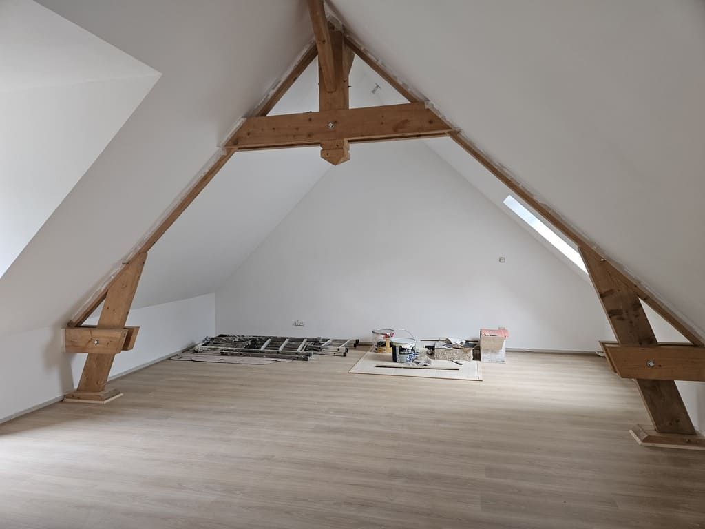 Intérieur de grenier avec poutres apparentes, murs blancs et sol imitation bois. Des outils de chantier sont disposés au centre.