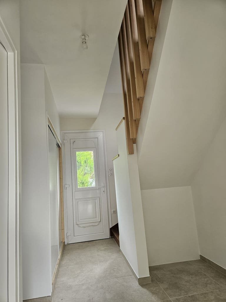 Couloir avec escalier et porte blanche ornée, rampe en bois naturel, murs blancs et sol gris.