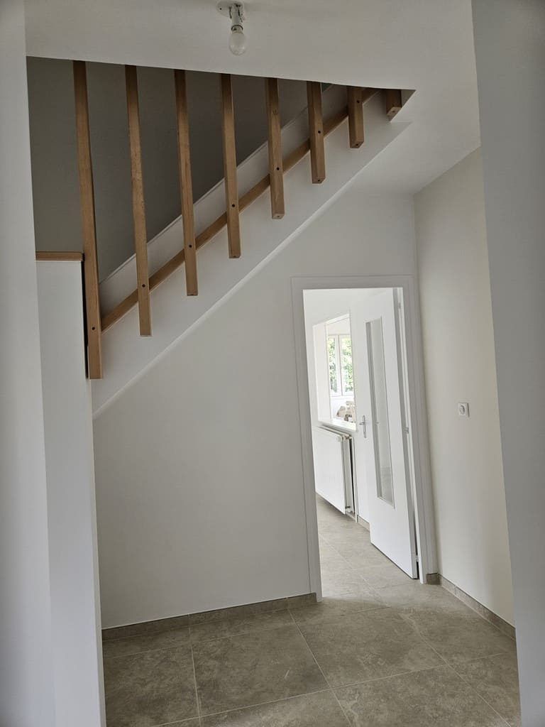Escalier avec rampe en bois, menant à un couloir aux murs blancs et à une porte ouverte.