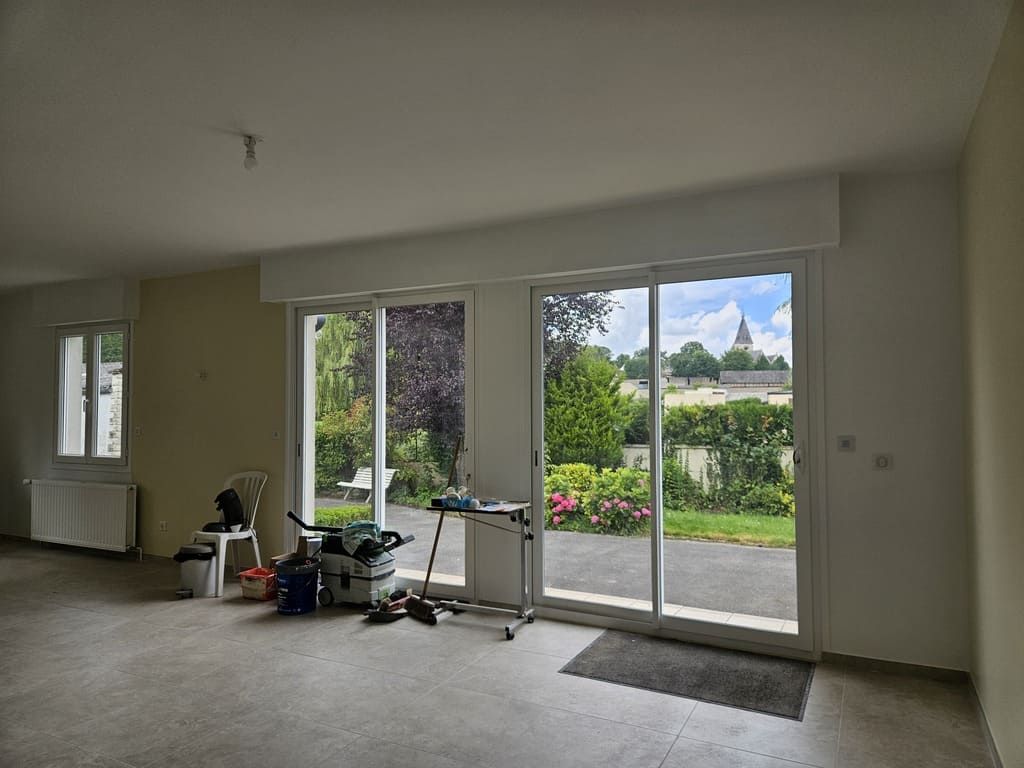 Pièce vide avec de grandes portes coulissantes en verre donnant sur un jardin et une église au loin.