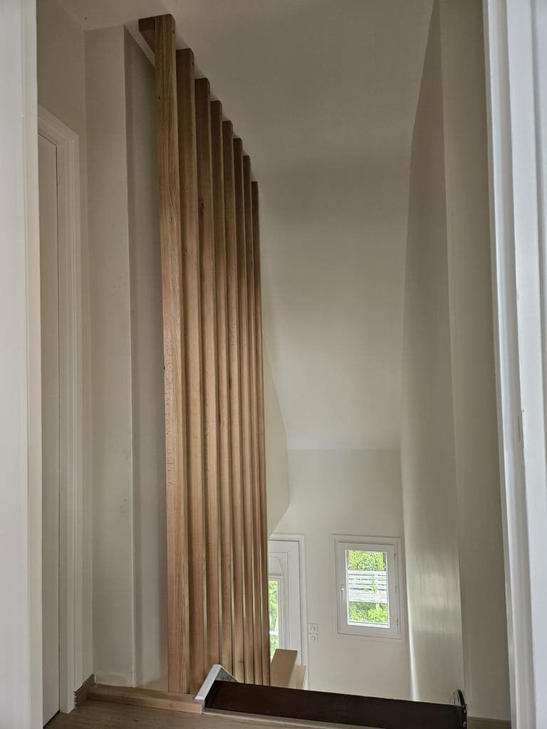 Cloison en lattes de bois séparant un escalier, tons de bois naturel sur murs blancs.