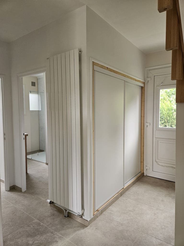 Couloir avec portes coulissantes blanches, radiateur et porte entrouverte donnant sur une salle de bains.