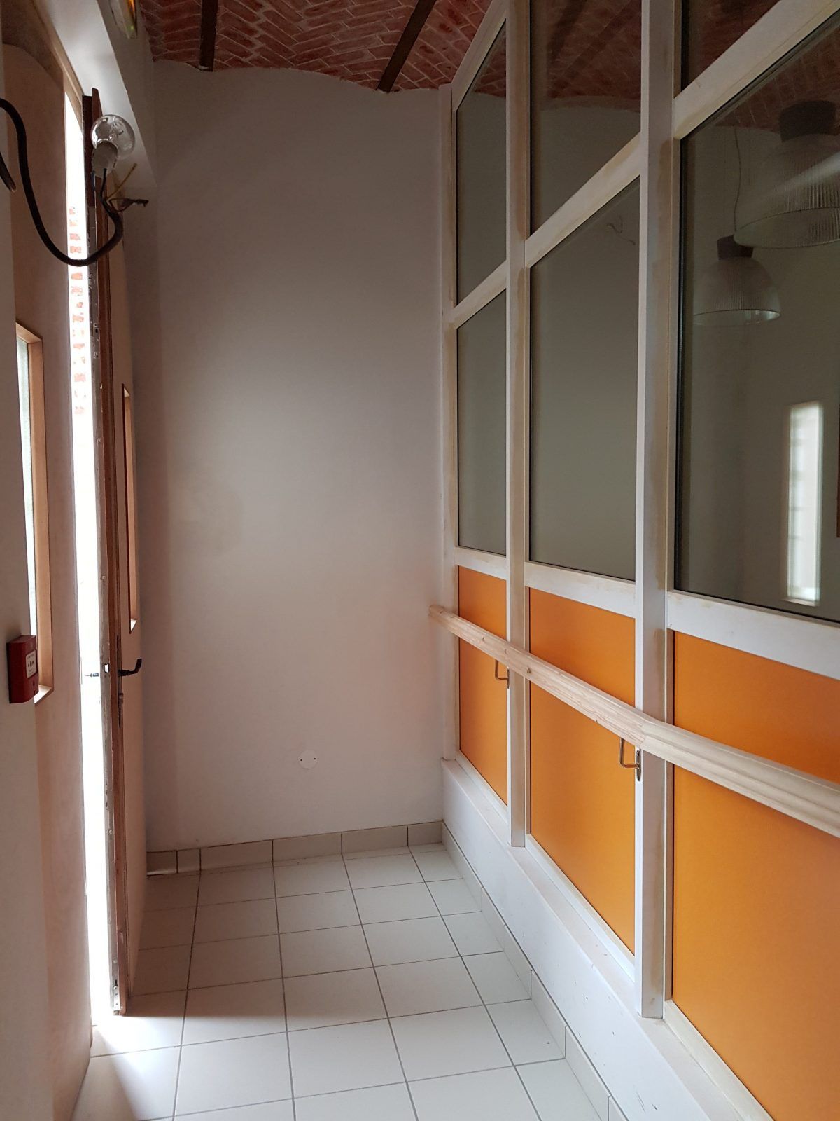 Couloir étroit aux murs blancs, lambris orange et grandes fenêtres.