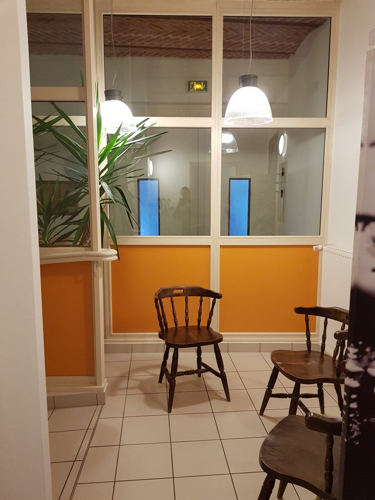 Salle d'attente avec chaises en bois, mur orange et fenêtres en verre.