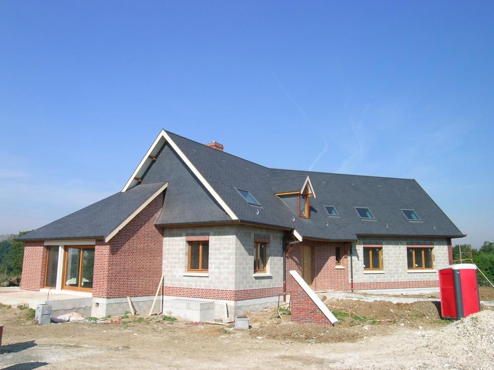 Maison en construction, façade en parpaings gris et briques rouges, toit gris foncé, ciel bleu.
