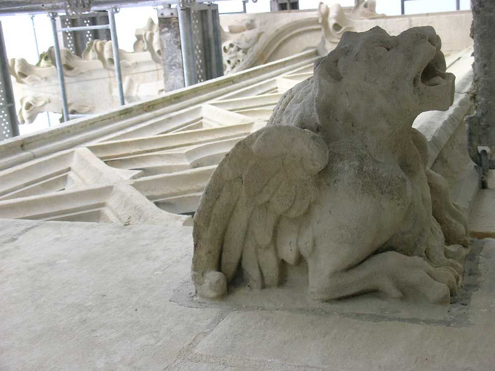 Statue de créature ailée en pierre sur le toit d'un bâtiment, gueule ouverte, ailes déployées.