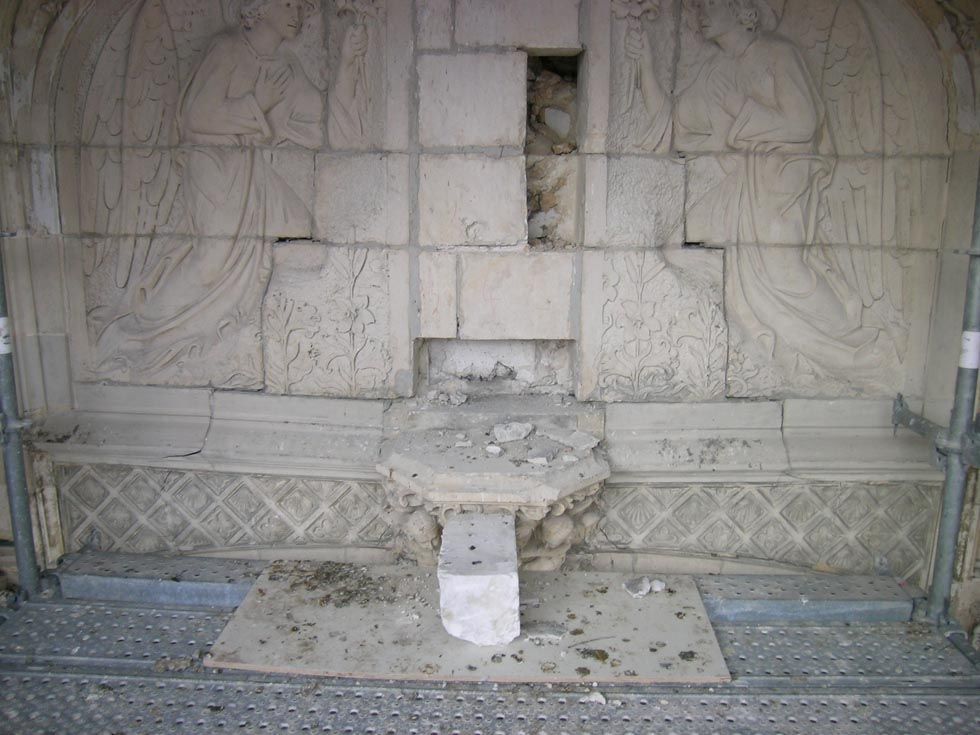 Mur en pierre orné d'anges sculptés, endommagé au milieu, débris sur une surface métallique.