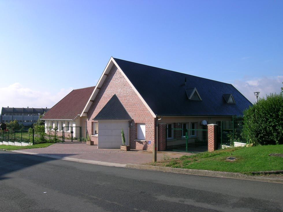 Maison à deux étages avec un toit sombre et un revêtement en briques, une allée et une petite cour verdoyante sous un ciel bleu.