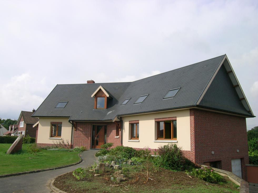 Maison avec toit gris foncé, façade en briques et bardage beige, fenêtres marron et allée carrossable.