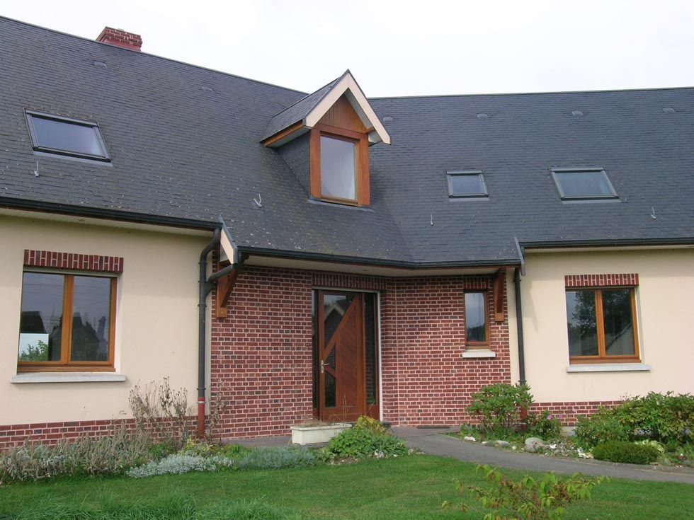 Maison de couleur beige avec toit foncé, entrée en briques et fenêtres en bois.
