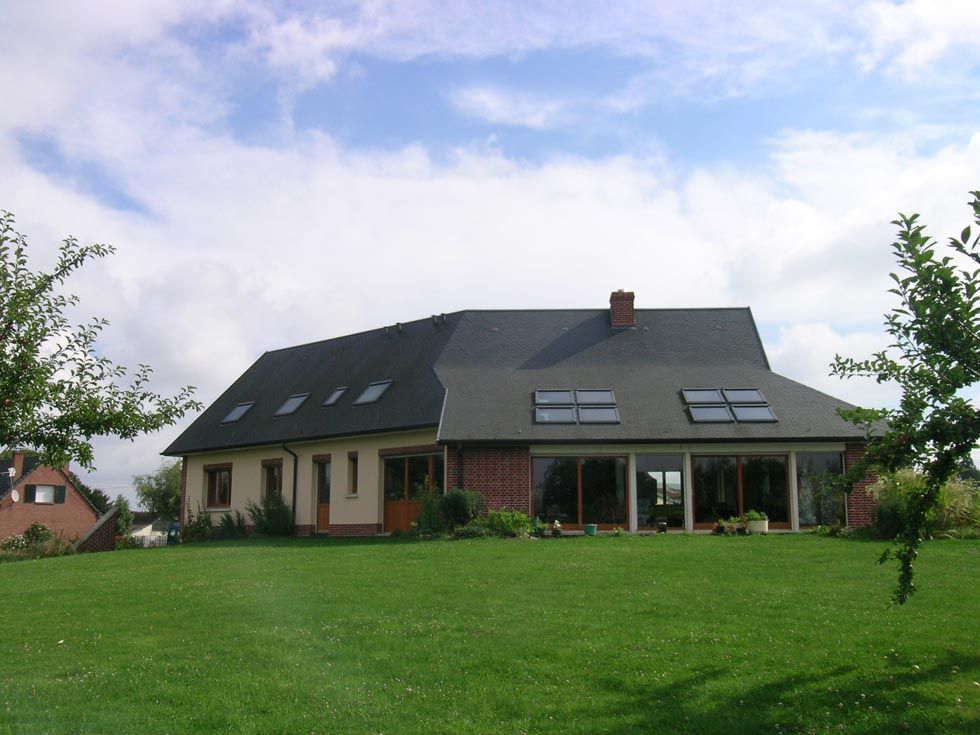 Maison avec de grandes fenêtres, des panneaux solaires et une pelouse verte sous un ciel bleu nuageux.
