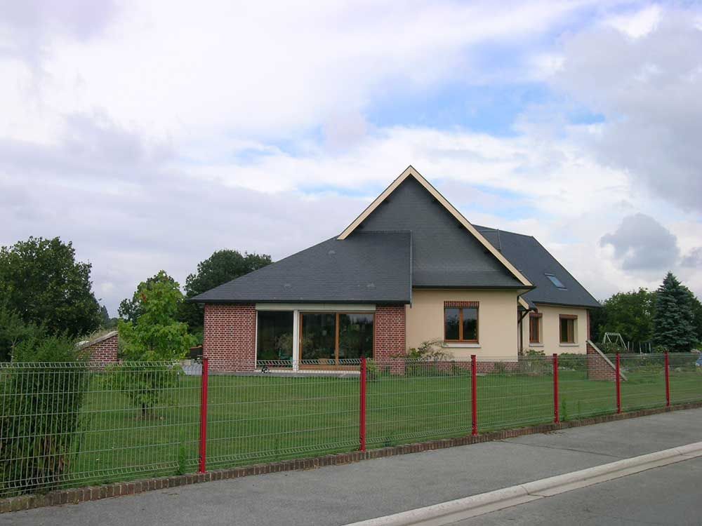 Une maison claire au toit sombre se cache derrière une cour verdoyante et une clôture à sommet rouge. Ciel nuageux.