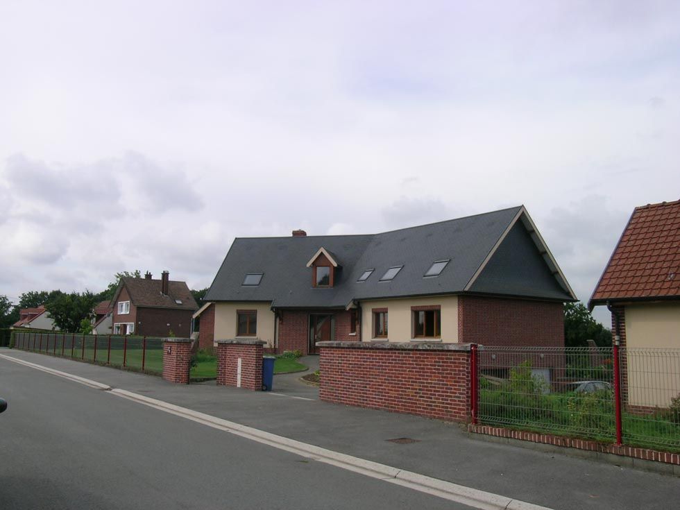 Une maison au toit d'ardoise, à la façade en briques beiges et à la clôture en briques et en métal. Ciel nuageux.