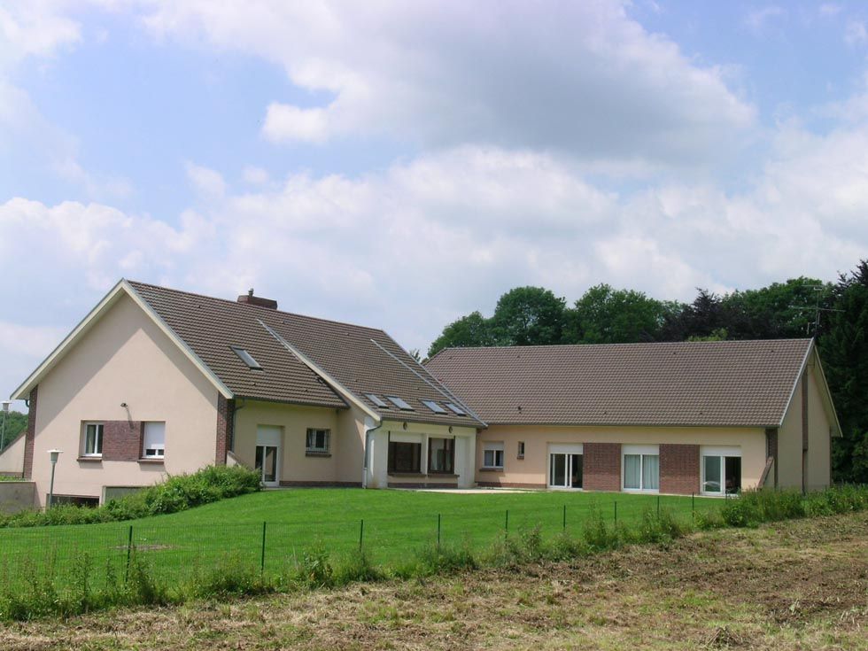 Maison beige au toit marron et à la pelouse verte, se détachant sur un ciel bleu nuageux.