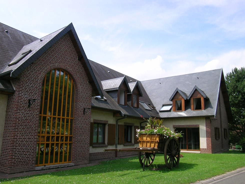 Bâtiment en briques avec une grande fenêtre cintrée, toit gris, volets en bois et une charrette à fleurs sur une pelouse verte.
