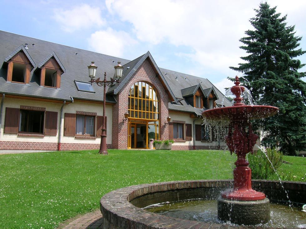 Bâtiment avec une fontaine au premier plan, pelouse. L'eau jaillit d'une fontaine rouge-brun. Ciel couvert.