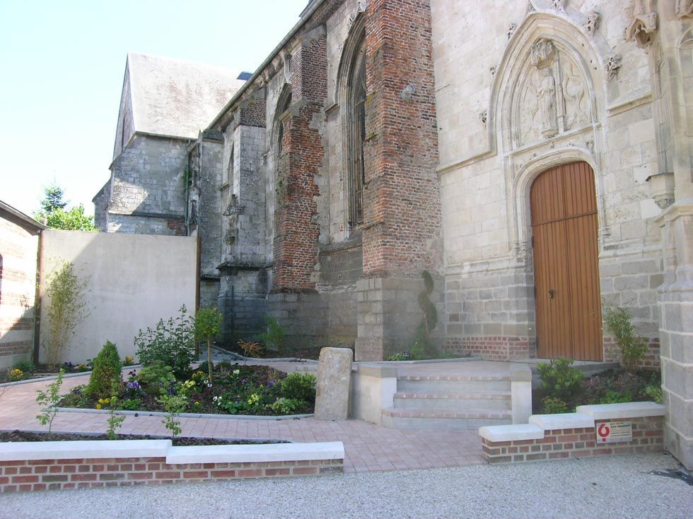 Vue latérale d'une église en pierre patinée par le temps, avec une porte en bois, entourée de briques et d'un petit jardin.