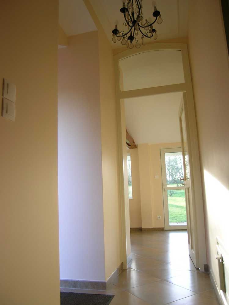 Couloir aux murs couleur pêche et orné d'un lustre, menant à une pièce avec une fenêtre et une porte donnant sur l'extérieur.