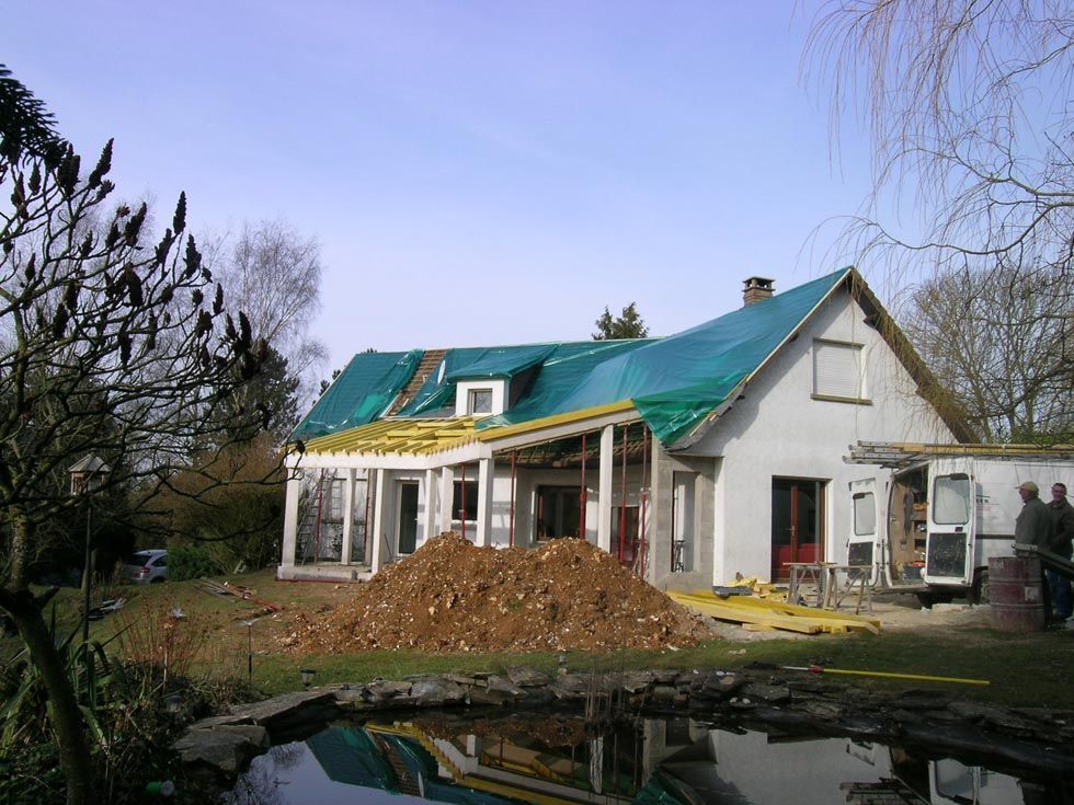 Maison en construction ; toit recouvert d’une bâche. Tas de terre, étang au premier plan, personnes visibles.