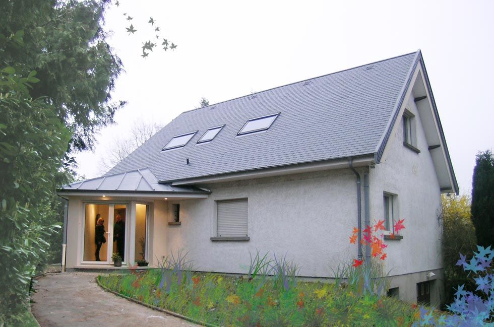 Maison blanche au toit gris en pente, avec puits de lumière et entrée couverte. Un chemin mène à la porte.