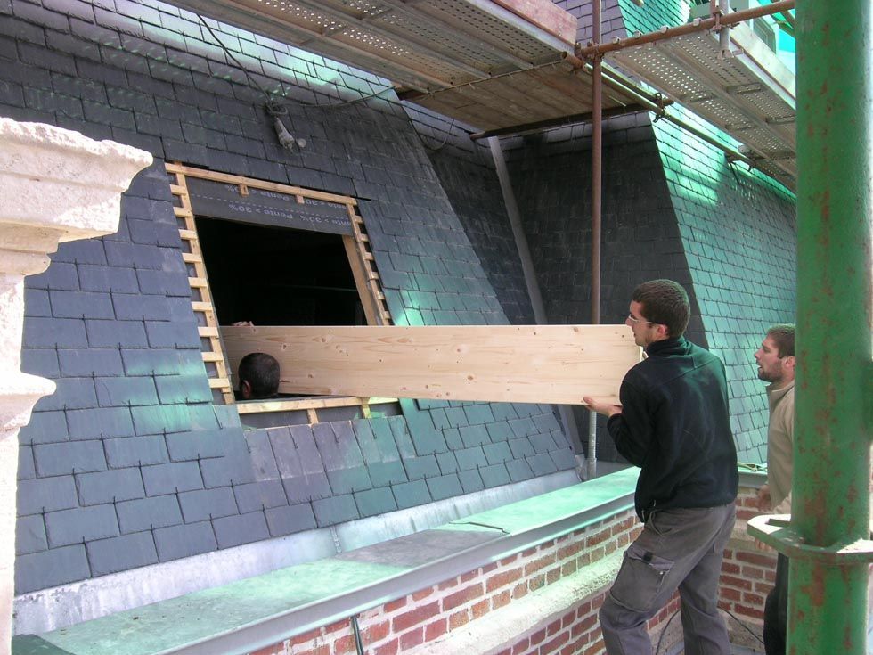 Deux hommes transportant une planche de bois près d'une ouverture dans le toit ; échafaudages et tuiles d'ardoise visibles.